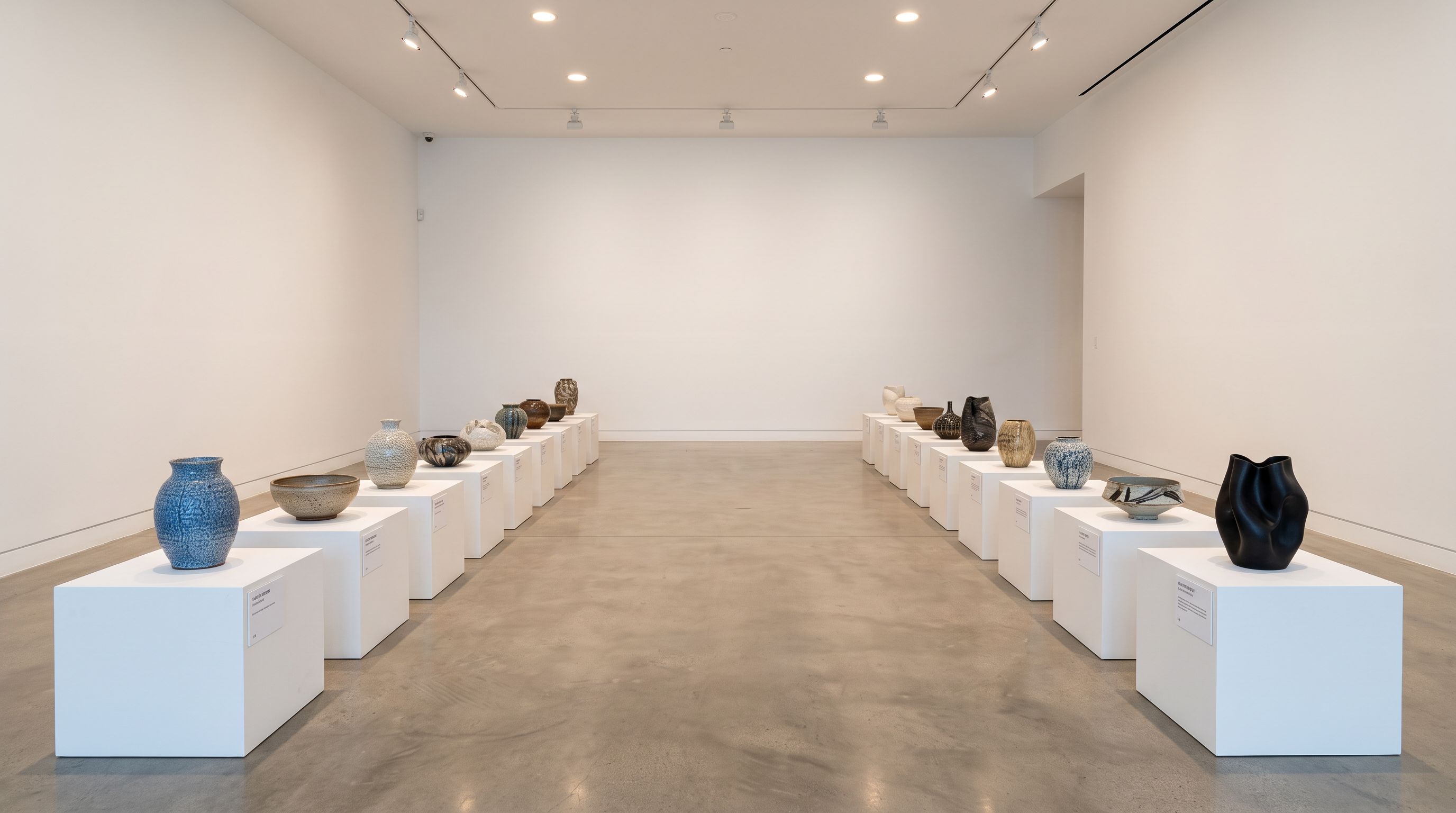 White-walled exhibition room with ceramic pieces on individual display plinths arranged in rows for a preview event, each with a label card under recessed spotlights.