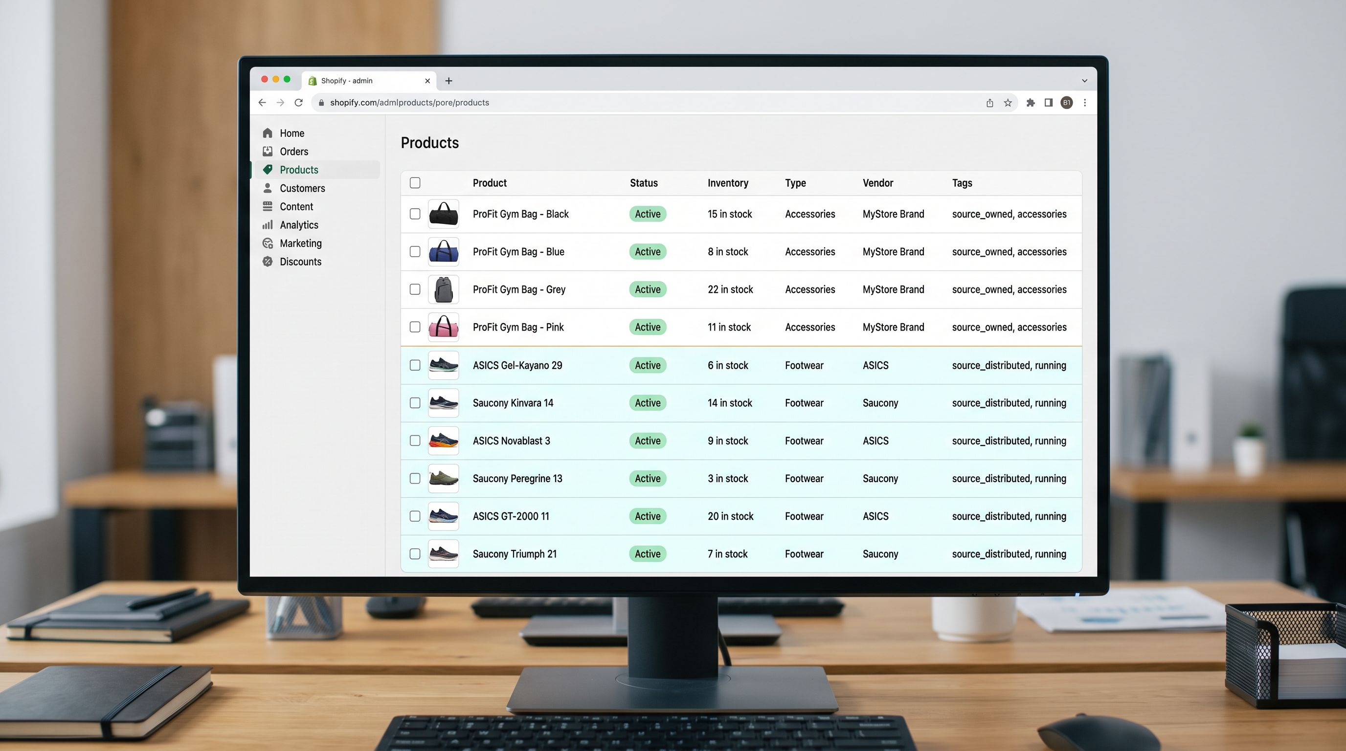 A Shopify admin product list on a monitor showing two groups of products: the top group showing four gym bag products with the Vendor column showing the store's own brand name, the bottom group showing six running shoe products with Vendor showing ASICS and Saucony, both groups correctly tagged in the Tags column with source owned and source distributed respectively, demonstrating correct catalogue separation after a structured mixed import, everything in sharp focus no blur no depth of field, colour photography.
