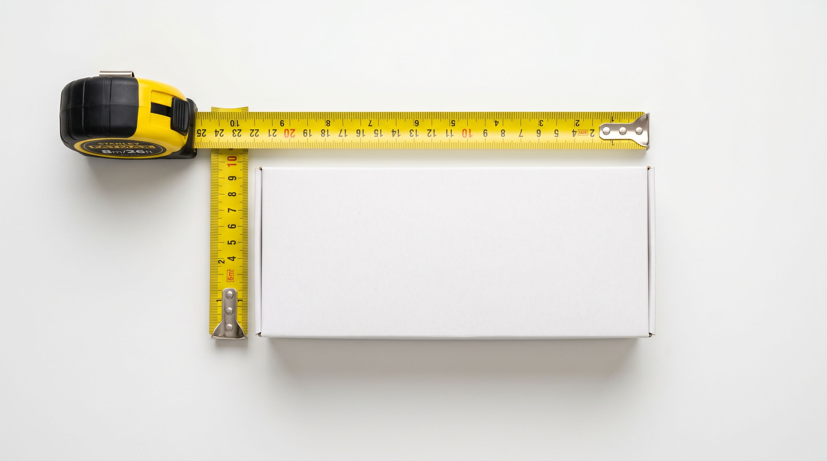 Yellow steel tape measure extended alongside a plain white product package on a white surface.