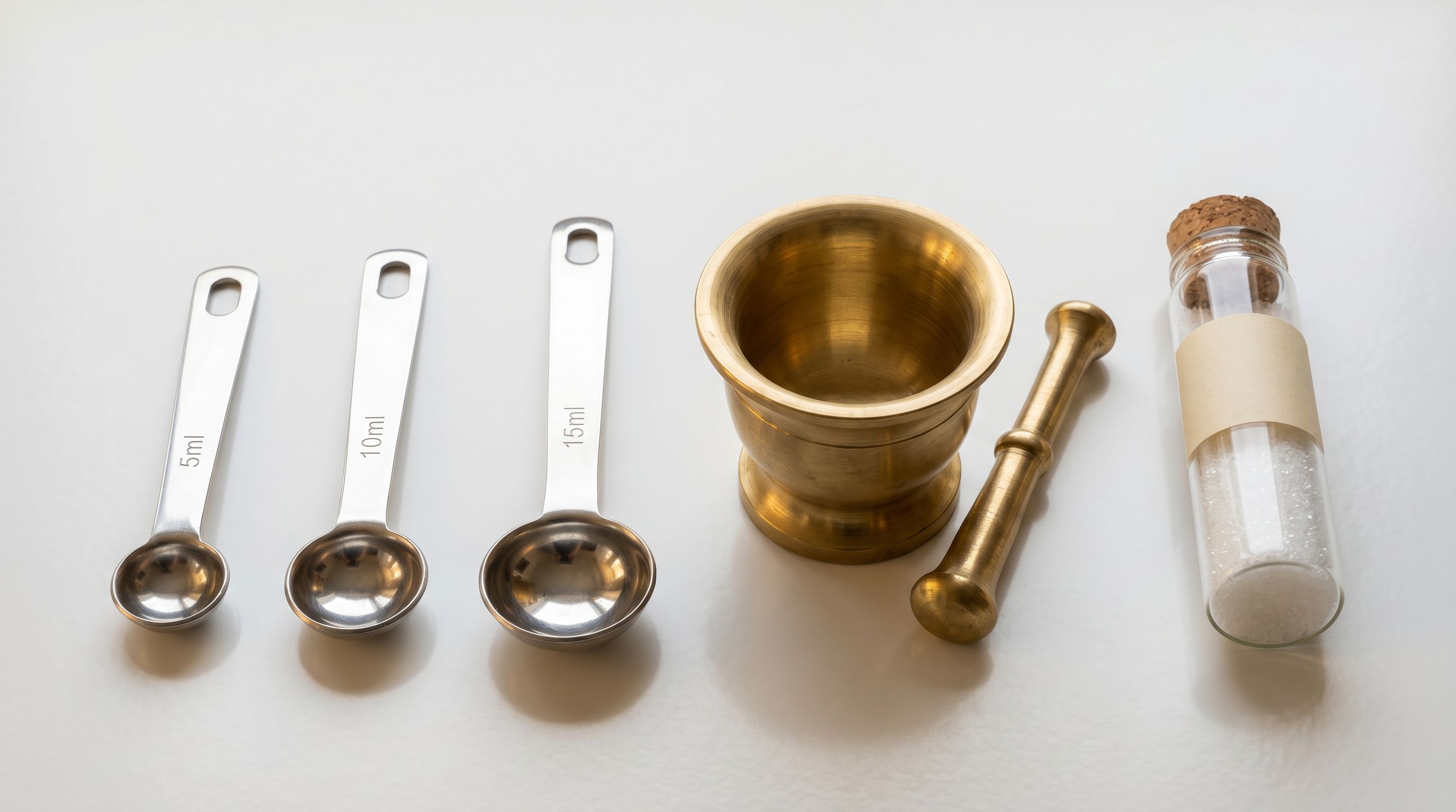 Precision measurement tools including calibrated spoons, a brass mortar and pestle, and a sealed glass vial with fine white granules on a white ceramic surface.