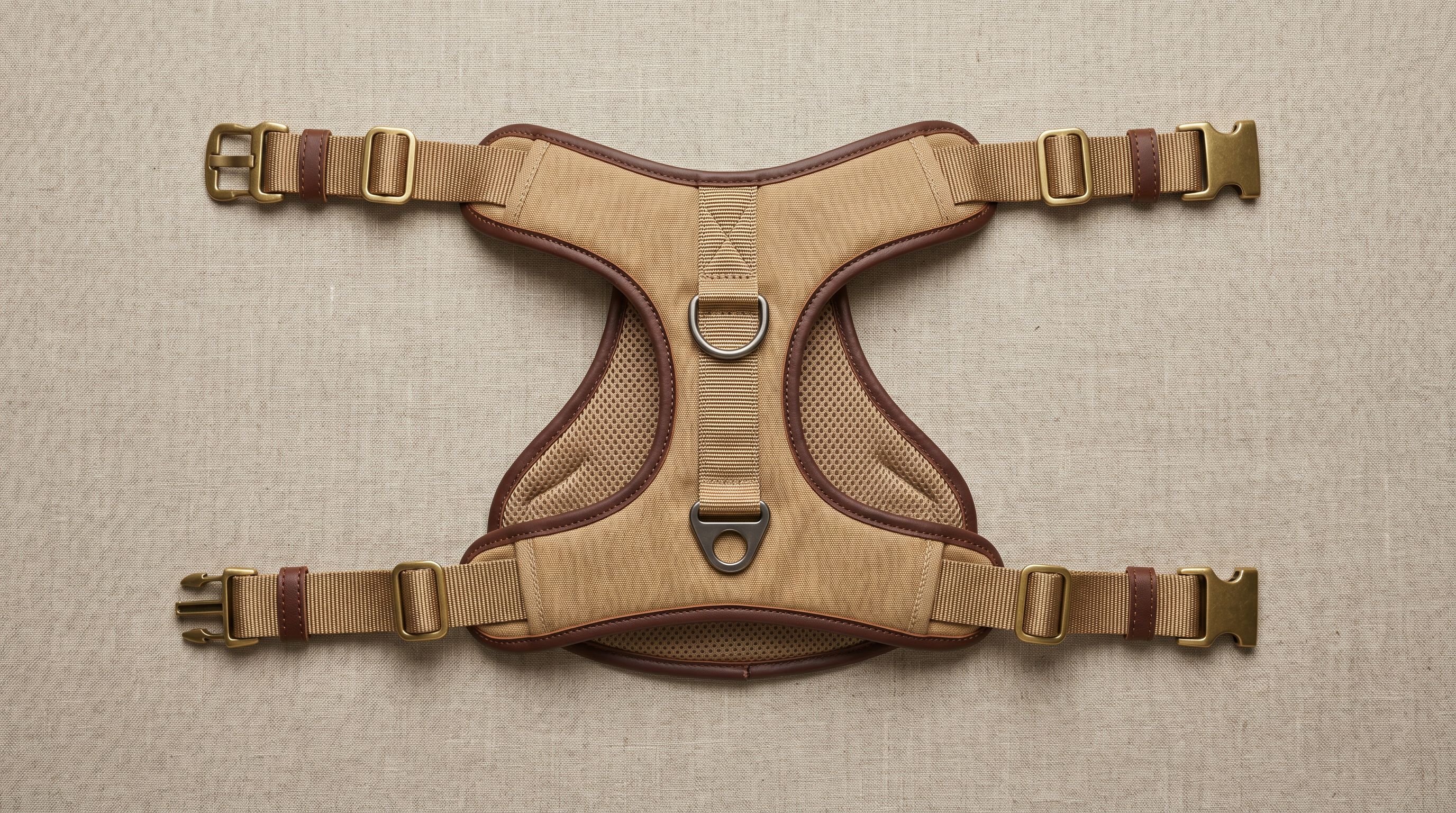 Structured tan canvas dog harness with leather trim laid flat on linen, showing padded chest plate, buckles, and front-clip and back-clip steel D-ring hardware.