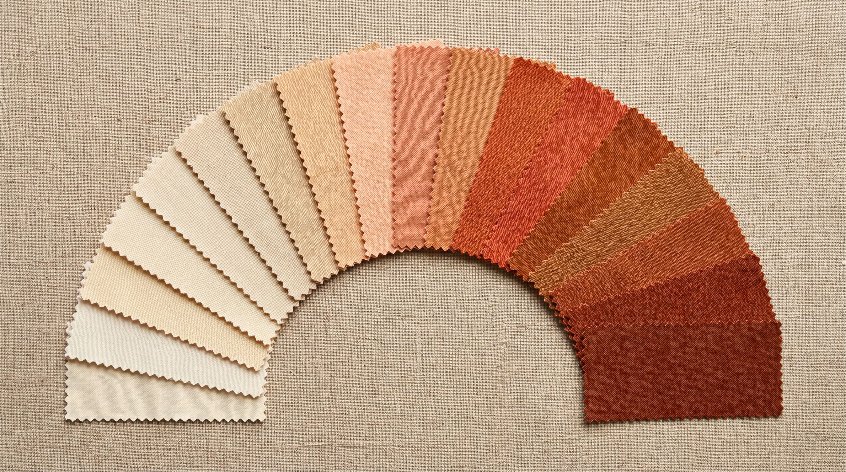 Fabric swatches in a graduated cream to rust colour spectrum fanned out on a linen surface.