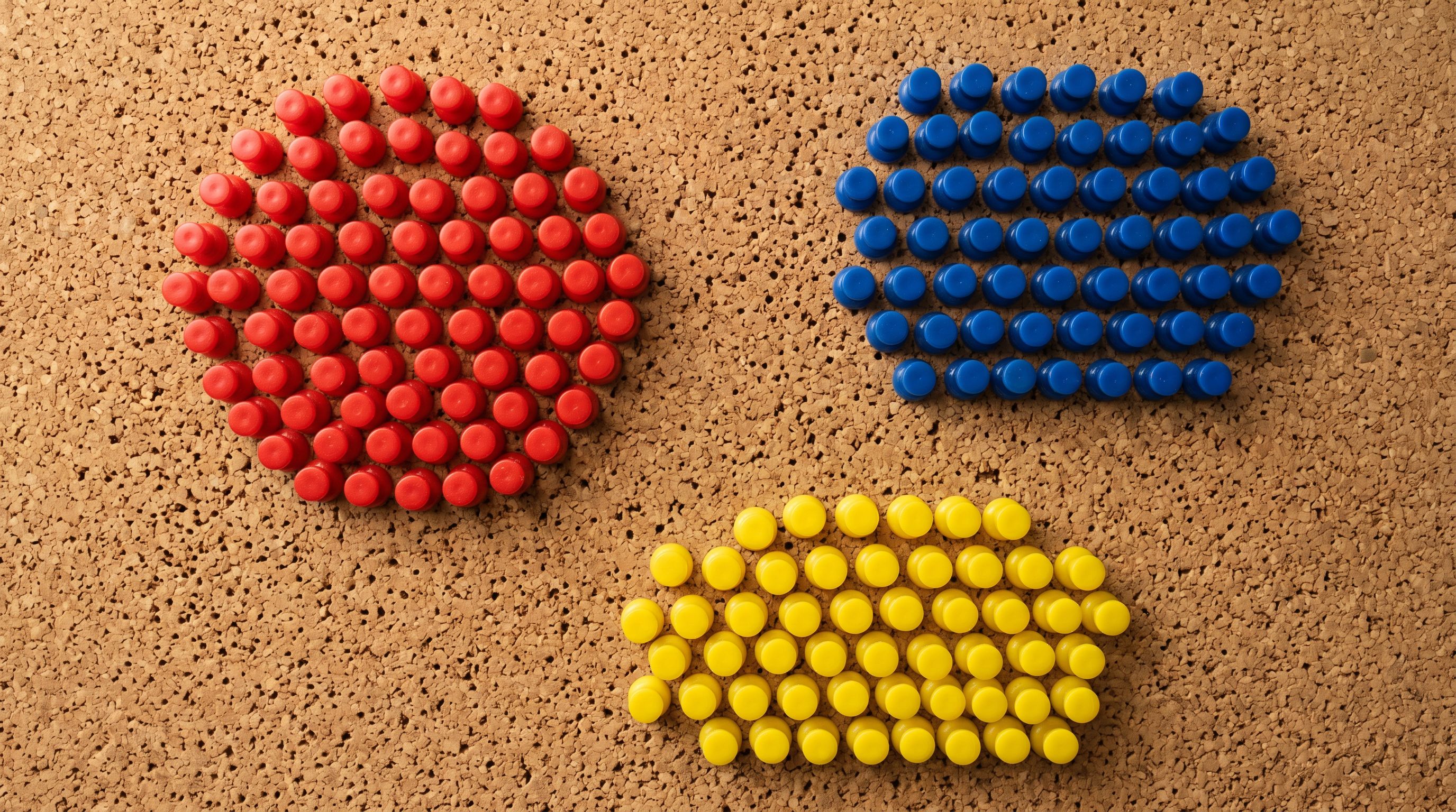 Colour-coded push pins clustered in distinct colour groups on a natural cork board surface.